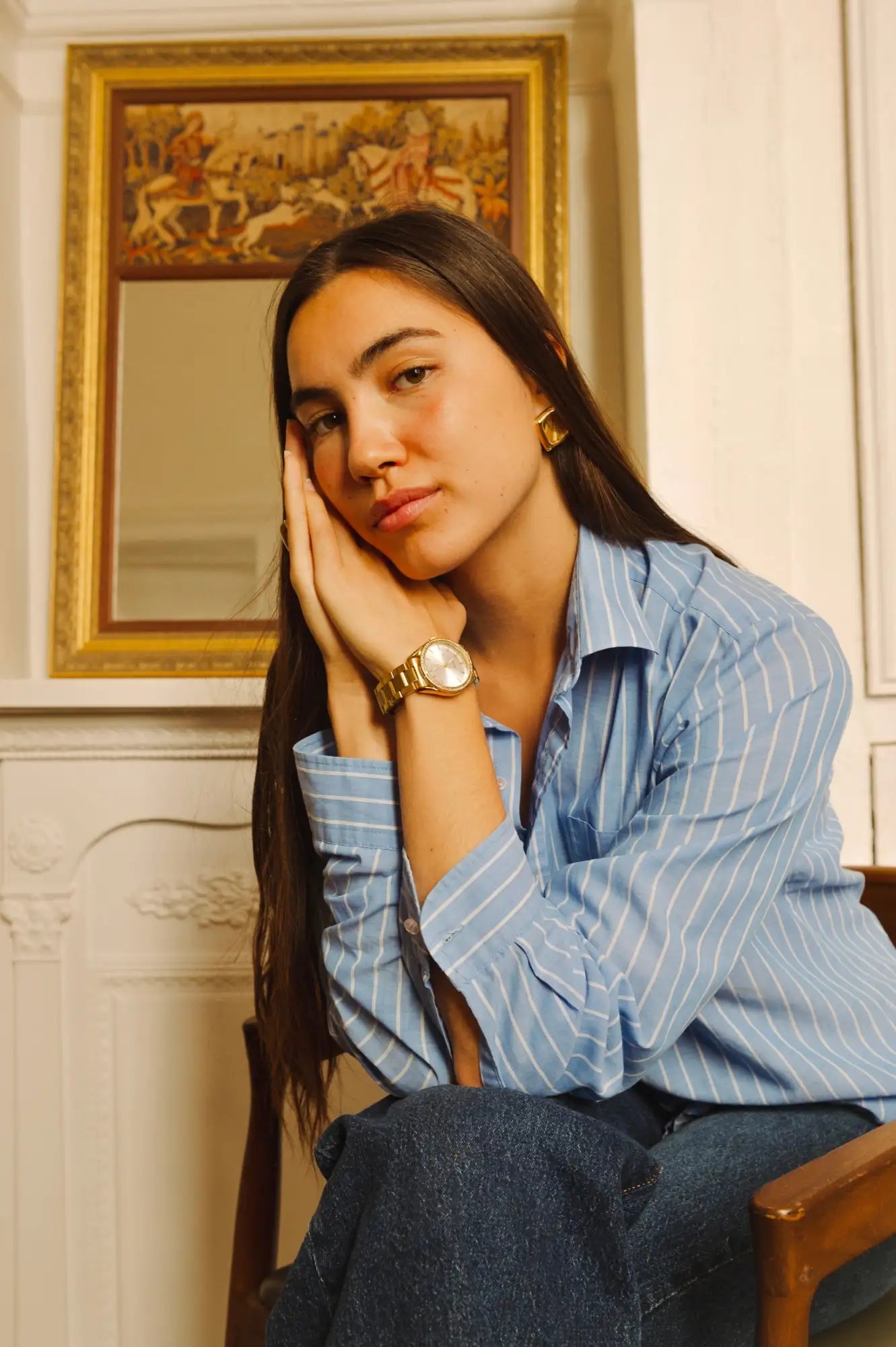 Montre à cadran rond et bracelet métallique de ton or portée au poignet gauche de la femme.